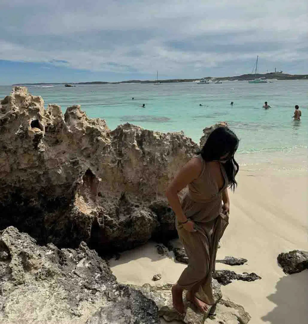 A woman in a flowing beige dress standing on a rocky terrain by the beach, with turquoise water and distant boats in the background.