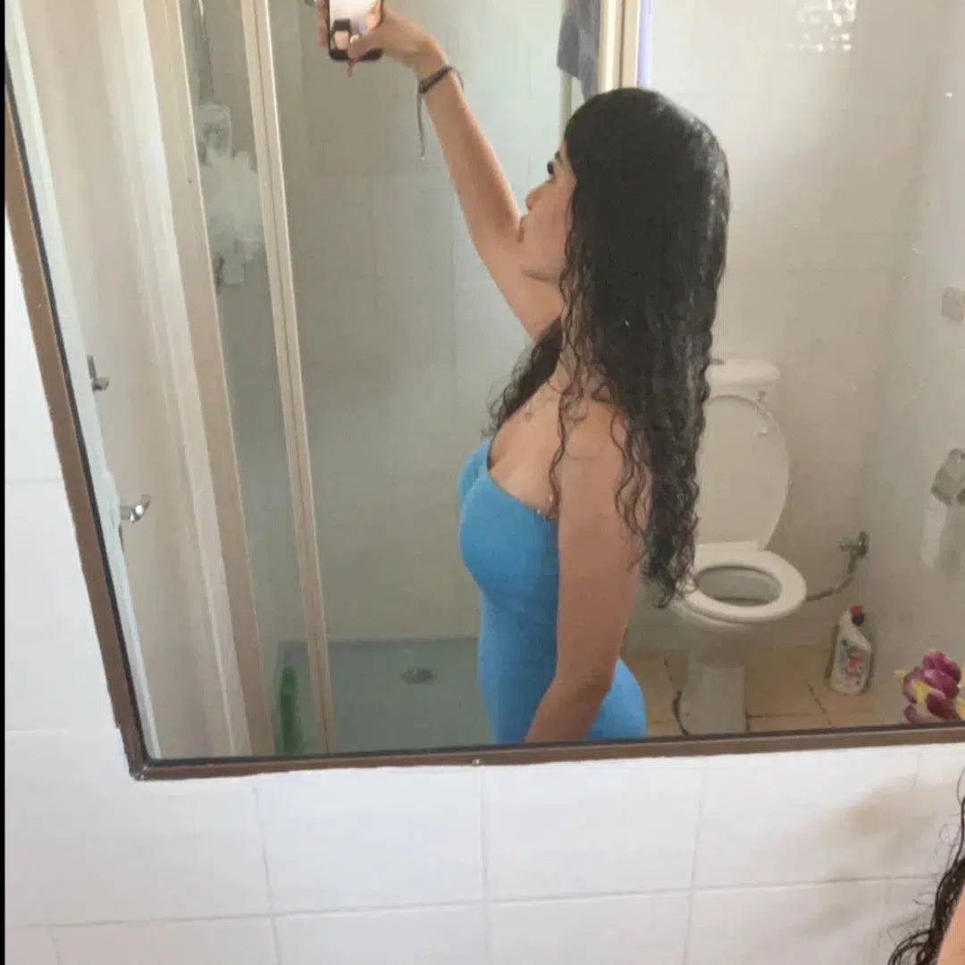 Mirror selfie of a girl with long curly hair wearing a blue dress in a bathroom setting.